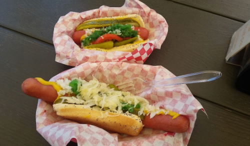 Two hot dogs in paper baskets: one topped with mustard, sauerkraut, and green onions; the other with relish and pickles.