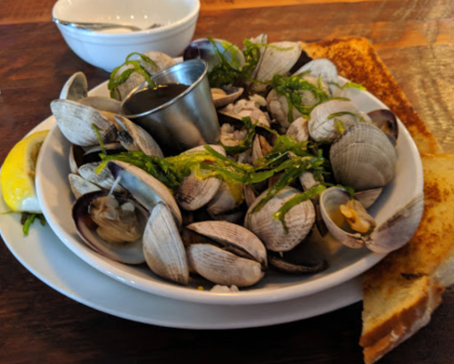 A plate of steamed clams garnished with seaweed, lemon, and a small cup of sauce, served with toasted bread.