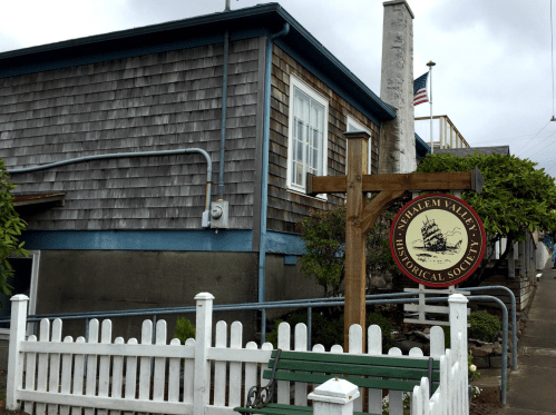 Sign for the Nahcotta Valley Historical Society in front of a wooden building, with a white picket fence and greenery.