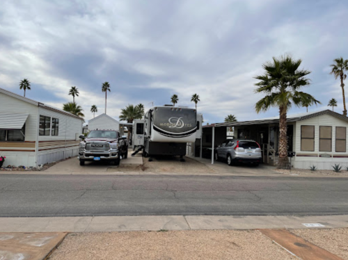 A parked RV between two mobile homes, with palm trees lining the street under a cloudy sky.