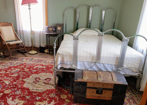 A vintage bedroom featuring a metal bed, a wooden rocking chair, a decorative rug, and an antique trunk at the foot of the bed.