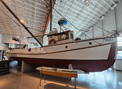 A vintage wooden boat named "Darle" displayed indoors, with a modern museum setting in the background.