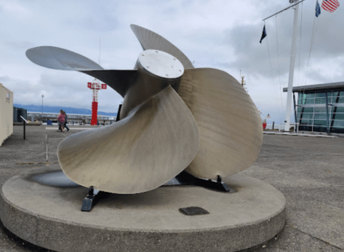 A large metallic ship propeller sculpture displayed on a concrete base near a waterfront building.