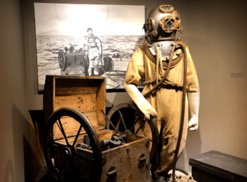 A vintage diving suit stands next to a wooden chest and a black-and-white photo of divers at sea.