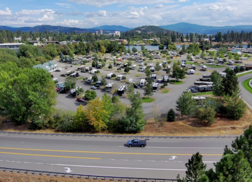 Aerial view of a large RV park with trees, a road, and mountains in the background under a partly cloudy sky.