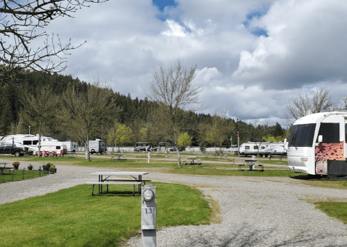 A scenic RV park with gravel paths, picnic tables, and trees, set against a backdrop of hills and cloudy skies.