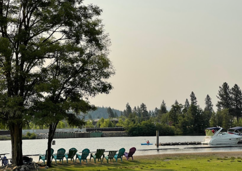A serene lakeside scene with colorful chairs, a boat dock, and a small boat on the water surrounded by trees.