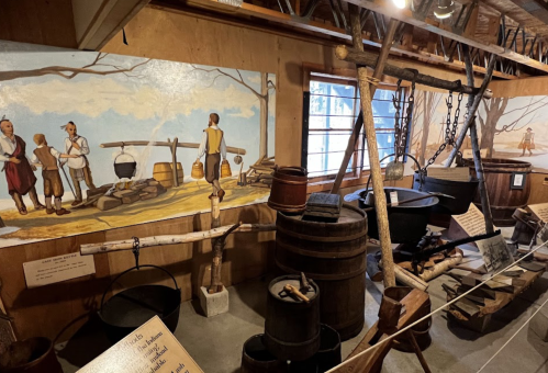 Interior of a historical exhibit featuring wooden barrels, cooking pots, and murals depicting early settlers.