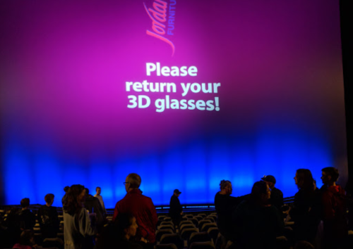 A theater interior with a colorful backdrop displaying "Please return your 3D glasses!" as people gather.