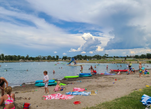 Children playing on a sandy beach by a lake, with colorful floats and a blue slide in the background under a cloudy sky.
