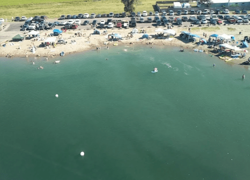 Aerial view of a beach area with people swimming, tents, and parked cars along the shore.