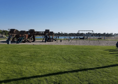 A playground with a wooden structure and swings, surrounded by grass and a lake, with people enjoying the area.