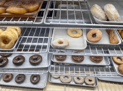 A display of various donuts on metal trays, including glazed, powdered, and chocolate varieties.