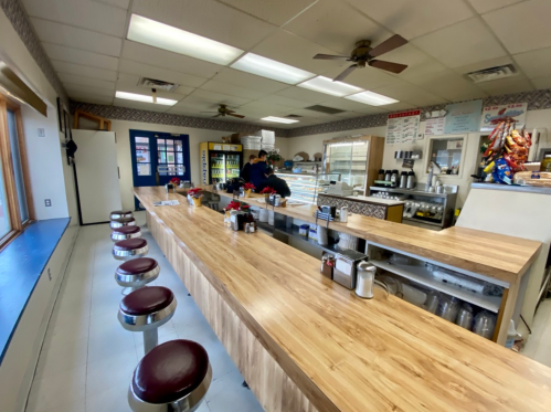 A cozy diner interior with a long wooden counter, stools, and a display case, featuring a staff member preparing food.