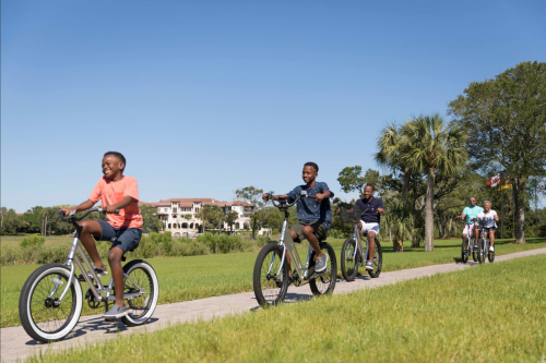 A group of boys joyfully riding bikes along a sunny path with greenery and a large house in the background.
