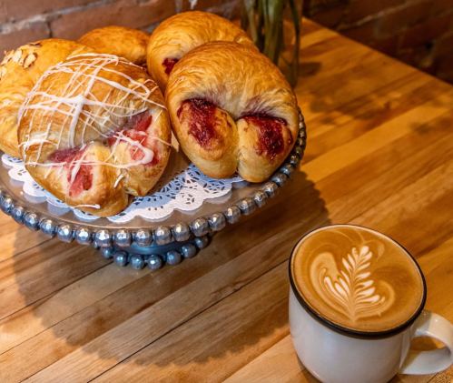 A plate of pastries with icing and jam, next to a cup of latte with a leaf design on a wooden table.