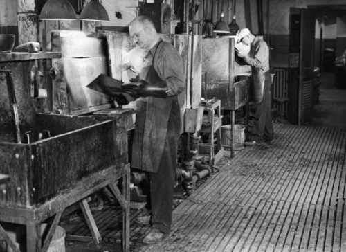 Two workers in a vintage industrial setting, using machinery and tools in a dimly lit workshop.