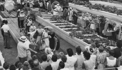 A crowd watches a festive event in a garden, featuring performers in costumes and decorated garden beds.