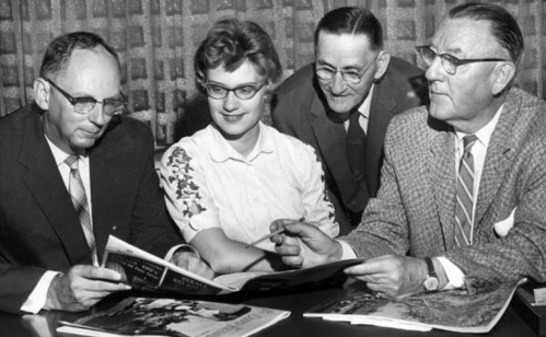 Four people, two men and two women, gather around a table, examining magazines and discussing.