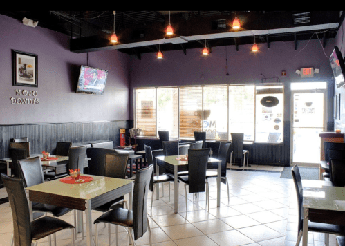 Interior of a donut shop with purple walls, tables, and chairs, featuring a TV and large windows for natural light.