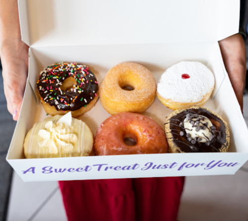 A person holds a box of six assorted donuts, featuring sprinkles, chocolate, and cream toppings, with a sweet message on the box.