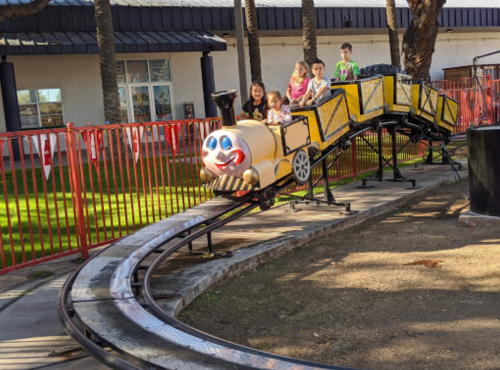 A small amusement park train ride with children smiling as it goes around a track in a sunny outdoor setting.