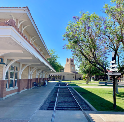 Historic train station with tracks, surrounded by greenery and trees under a clear blue sky.