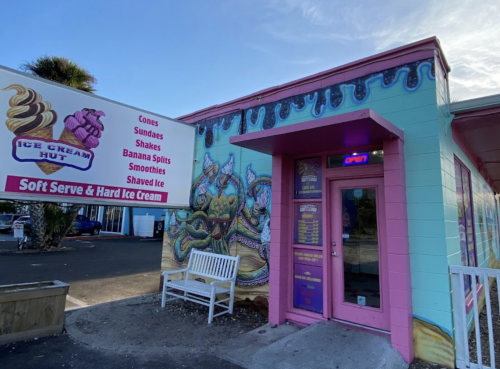Colorful ice cream shop exterior with a mural, sign listing treats, and a bench outside. Soft serve and hard ice cream available.