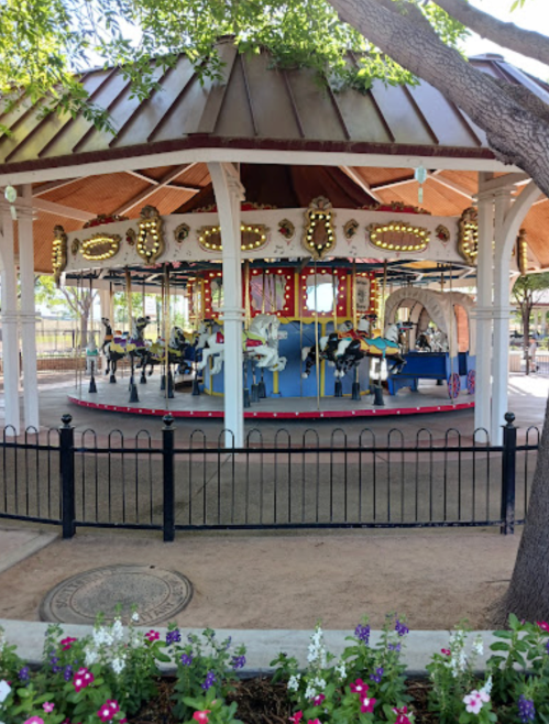 A colorful carousel with white horses, surrounded by a fence and blooming flowers, under a shaded pavilion.