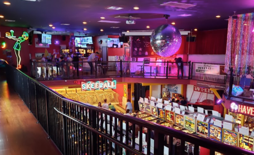 A vibrant arcade with skeeball, pinball machines, and colorful lights, featuring a disco ball and a lively atmosphere.
