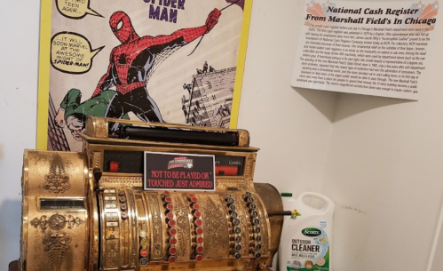 A vintage cash register with a Spider-Man poster above it, displayed alongside a cleaning product and an informational sign.