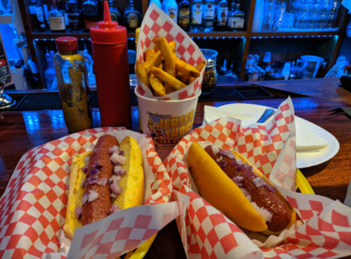 Two hot dogs with onions on a plate, accompanied by a cup of fries and condiments, set against a bar backdrop.