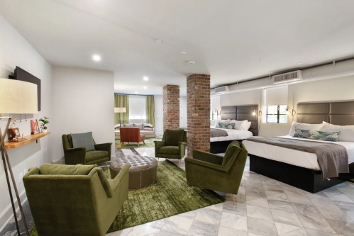 A modern hotel room featuring two beds, green armchairs, a coffee table, and exposed brick accents.