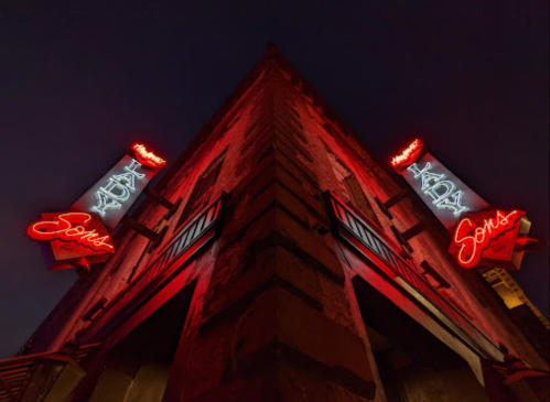 Neon signs for "Lady" and "Sons" illuminate a building corner at dusk, casting a warm red glow.