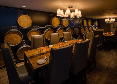 A dimly lit restaurant with a long wooden table, surrounded by leather chairs and wine barrels on the wall.
