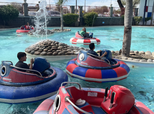 People enjoying bumper boats in a blue pool with a fountain and palm trees in the background.
