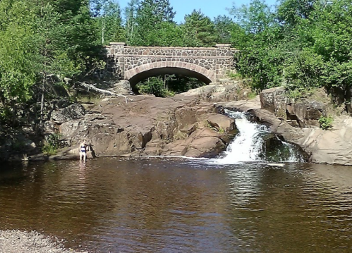 A stone bridge arches over a rocky area with a small waterfall, surrounded by lush greenery and a calm pool of water.