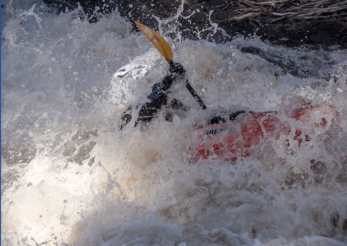 A kayaker navigates through turbulent whitewater, splashes of water flying around them.
