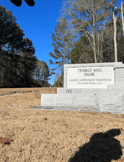 Sign for Tribble Mill Park with a clear blue sky and trees in the background, set on a grassy area.