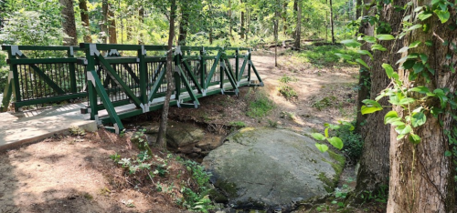A green wooden bridge spans a small creek in a lush, wooded area with trees and rocks surrounding it.