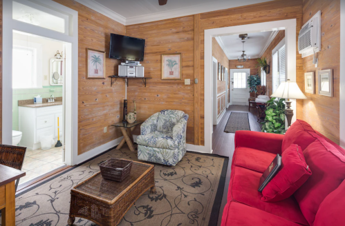 Cozy living area with wooden walls, a red couch, patterned chair, and a small TV, leading to a bright hallway.