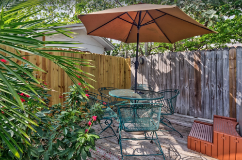 A cozy outdoor patio with a glass table, green chairs, an umbrella, and lush plants surrounded by a wooden fence.