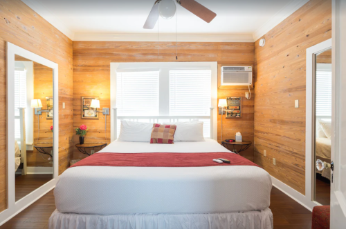 Cozy bedroom with wooden walls, a queen bed, mirrors, and a window air conditioner. Warm lighting and decor.