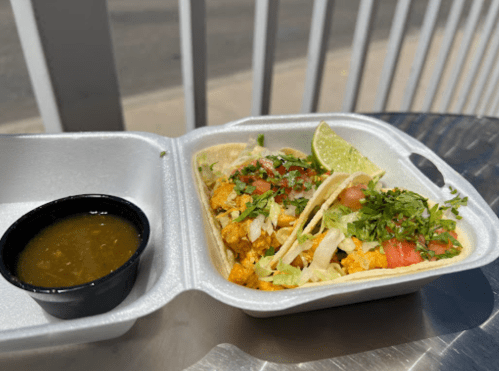 Two tacos with toppings and a lime wedge, served in a takeout container alongside a small cup of green salsa.