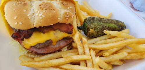 A sesame seed bun burger with cheese, bacon, lettuce, and a side of fries and a jalapeño pepper.