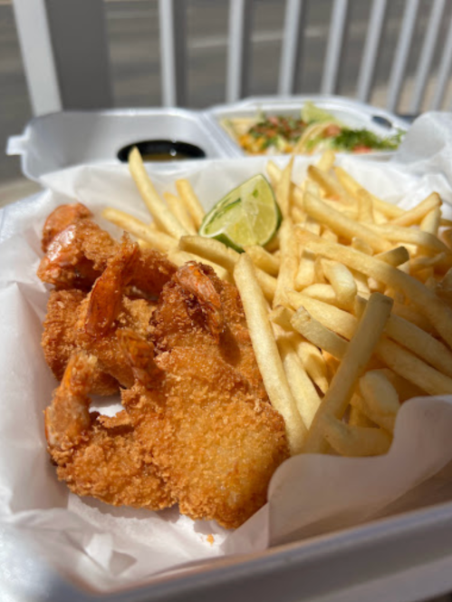 A takeout container with fried shrimp, French fries, lime, and dipping sauces on a table.