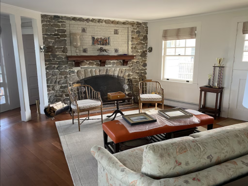 Cozy living room with a stone fireplace, two chairs, a coffee table, and a floral sofa, featuring natural light.