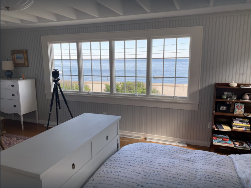 Cozy bedroom with a view of the beach through large windows, featuring a dresser, bookshelf, and a camera on a tripod.