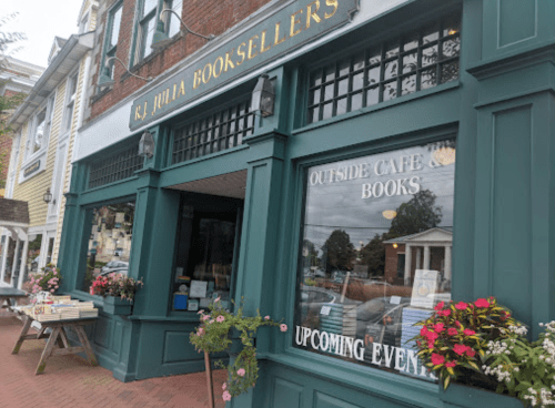 A charming bookstore exterior with a café, flowers, and a sign for upcoming events.
