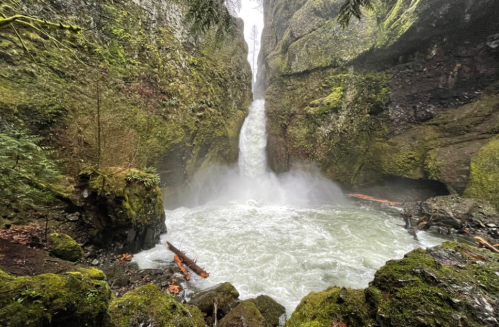 A waterfall cascades into a rocky pool surrounded by lush green moss and steep cliffs. Mist rises from the water.